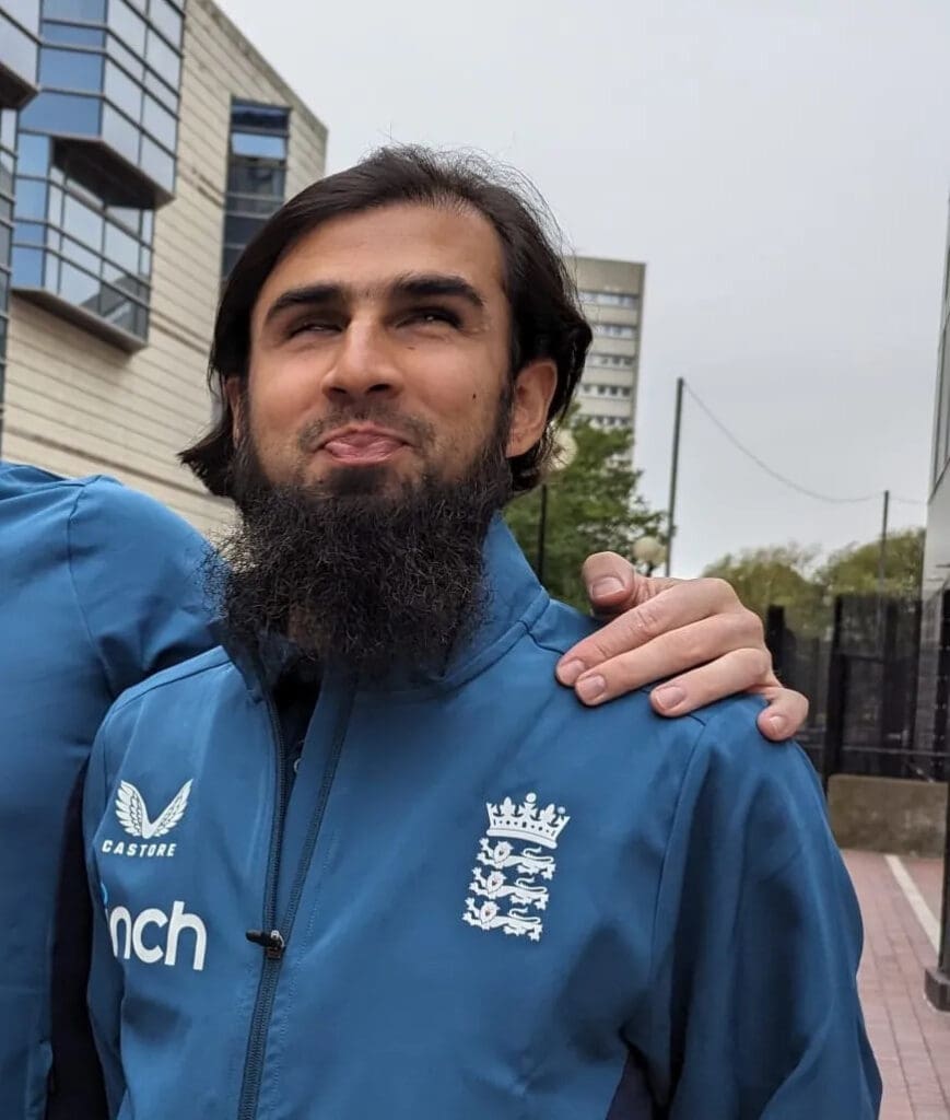 Mahomed smiles for a photo in a cricket uniform with someone who is out of frame with his hand on Mahomed's shoulder 