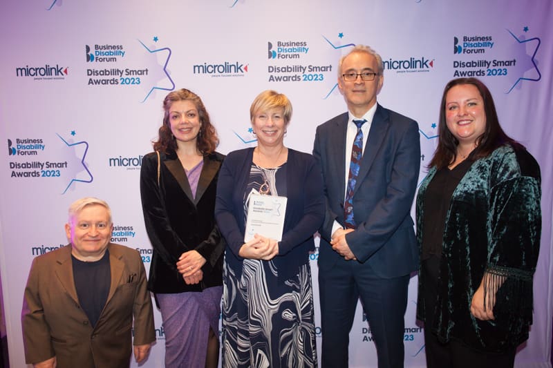 A group of people stand in from of a wall with Disability Smart Awards 2023 printed on it with Jane Hatton in the middle holding an award Evenbreak won 