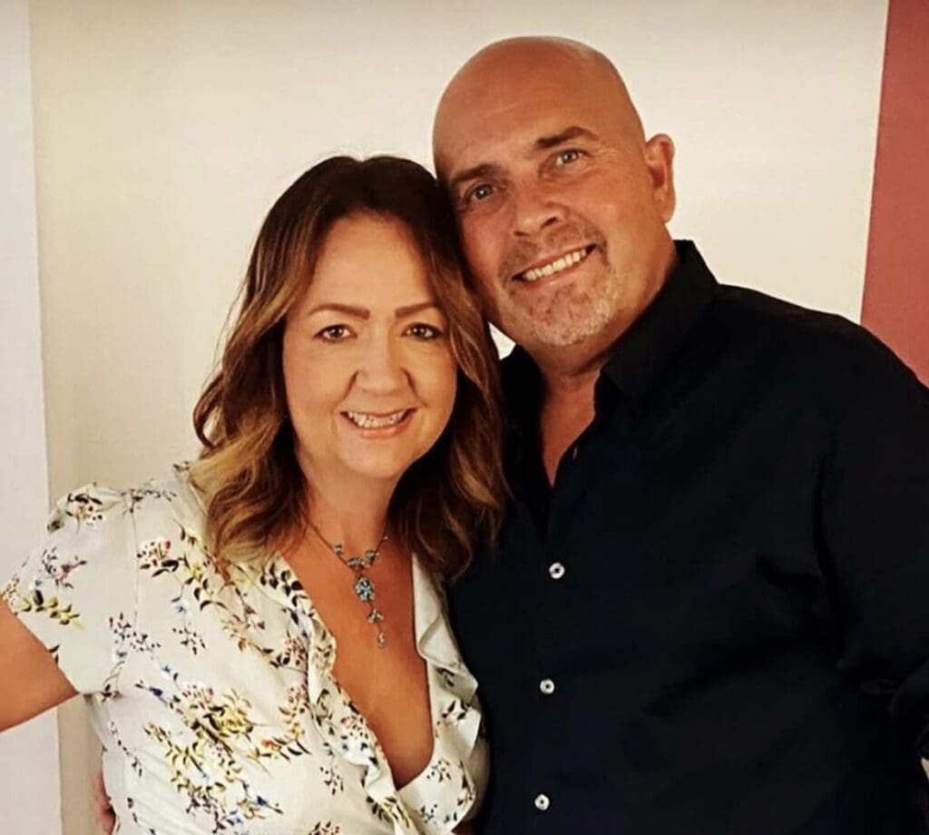 Nicola Glover on the right, wearing a floral dress with curled hair next to her partner Pete, wearing a black shirt . Both are smiling at the camera.
