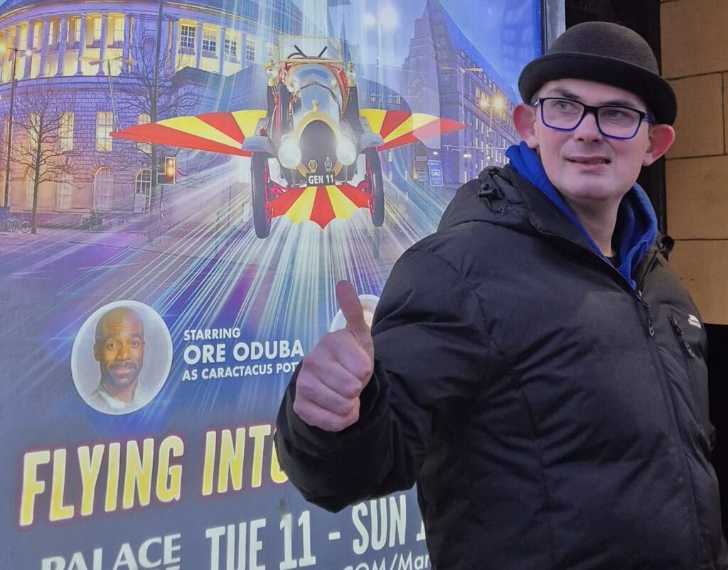 Zach in front of a movie poster, wearing a bowler hat and glasses, smiling with his thumb up to the camera.
